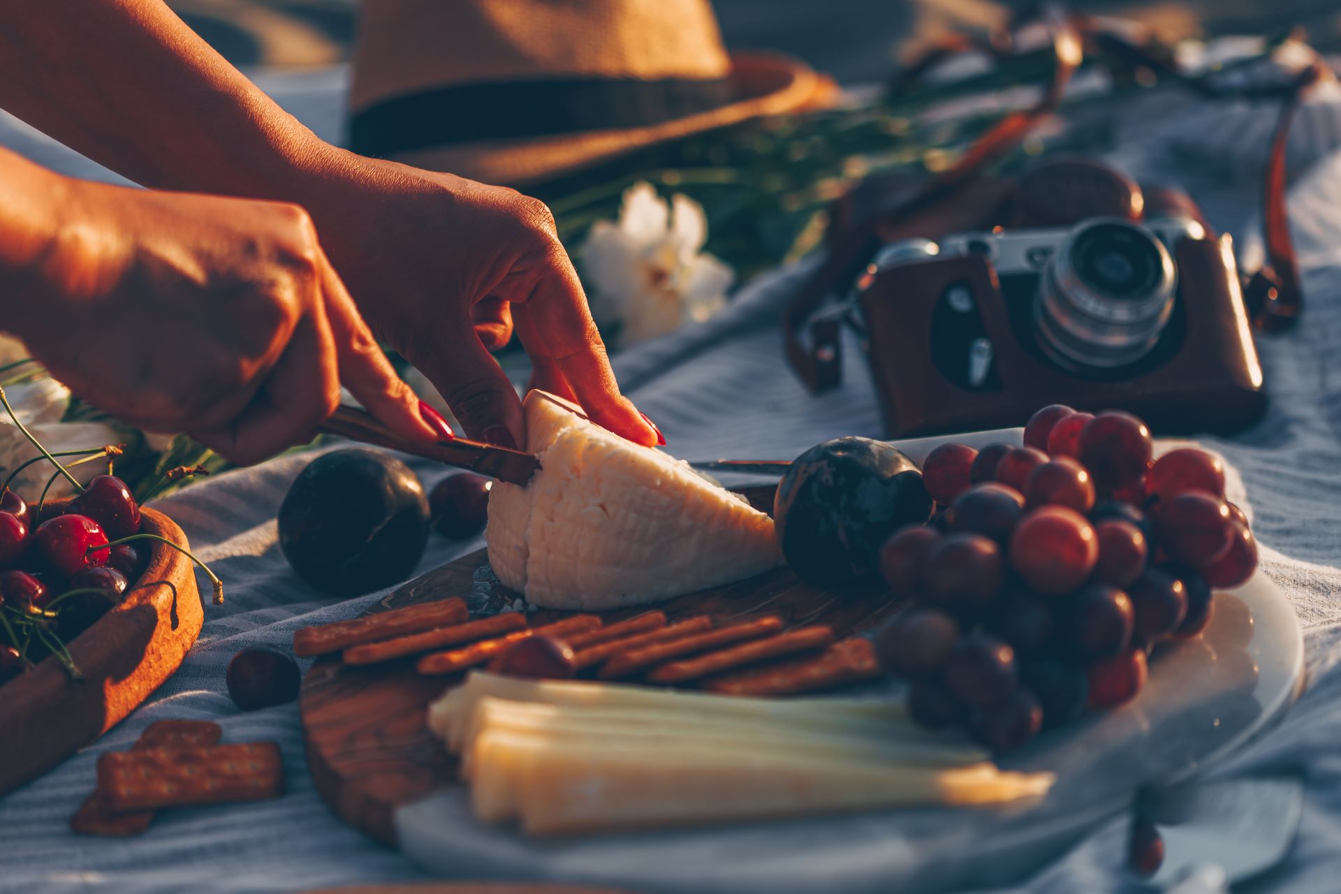 woman-slicing-cheese-wooden-cutting-board-with-cheese-fruits-it-camera-hat-flowers-beach
